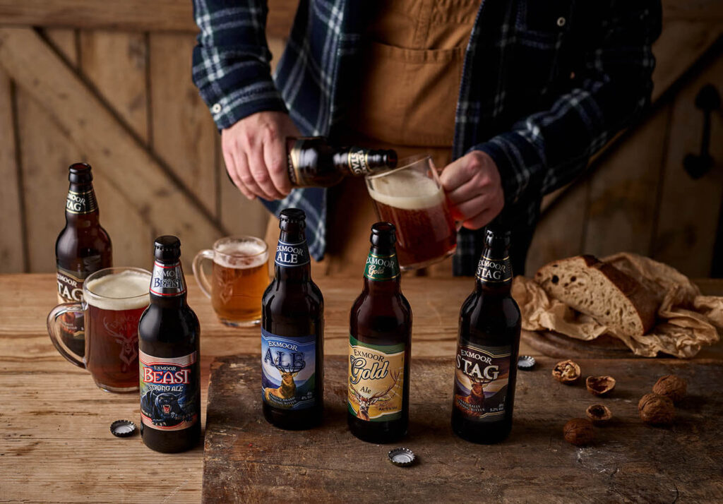 Man's hands pouring a beer into a pint glass with Exmoor Beast, Exmoor Ale, Exmoor Gold, and Exmoor Stag bottles on the table in front.