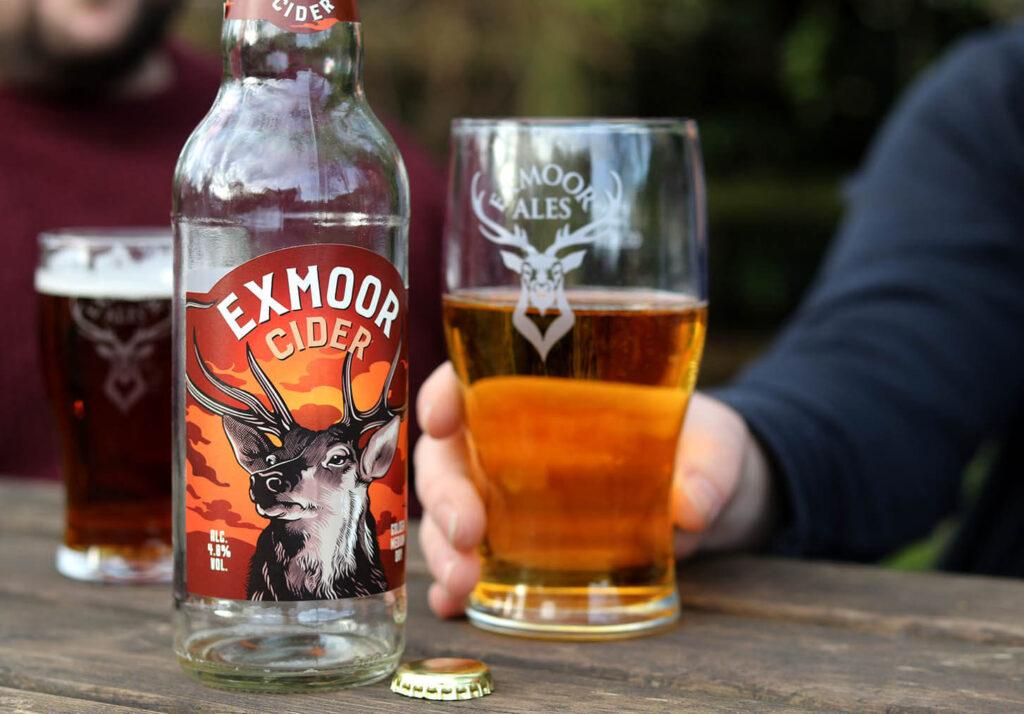 Exmoor Cider in a glass with a hand holding it and the bottle next to it sitting at a picnic table